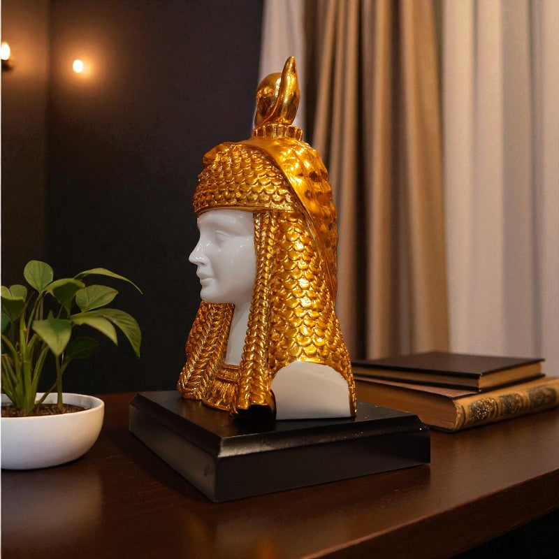 Decorative sculpture of a head with a gold headdress on a table with books and a plant.
