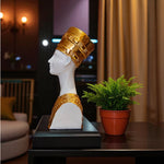 Statue of a head with gold headdress on a table with a plant in the background