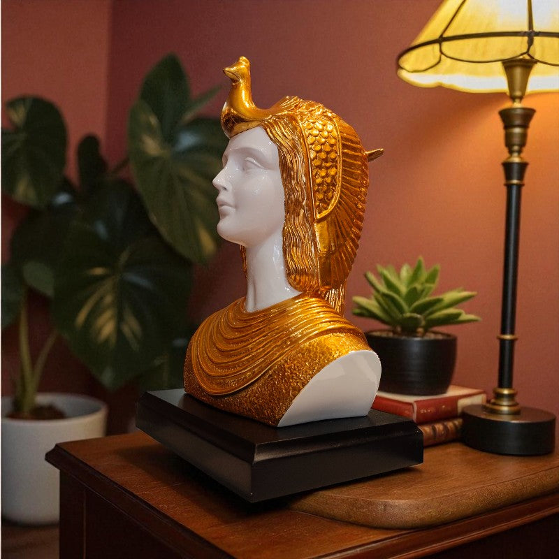 Decorative bust with gold and white finish on a wooden surface with plants and a lamp in the background.