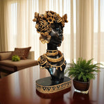 Decorative bust with floral and jewelry details on a table in a room with curtains and plants.