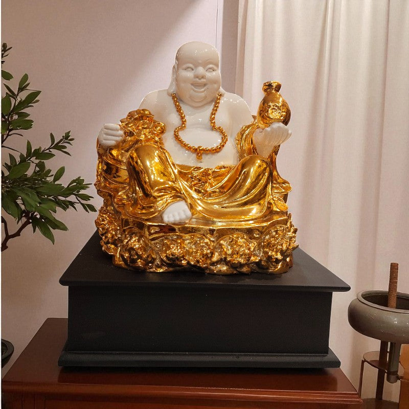 Gold and white Buddha statue on a black pedestal with a plant and curtain in the background