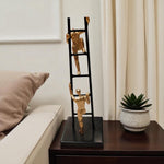 Decorative ladder sculpture with gold figures on a table next to a plant and pillow.