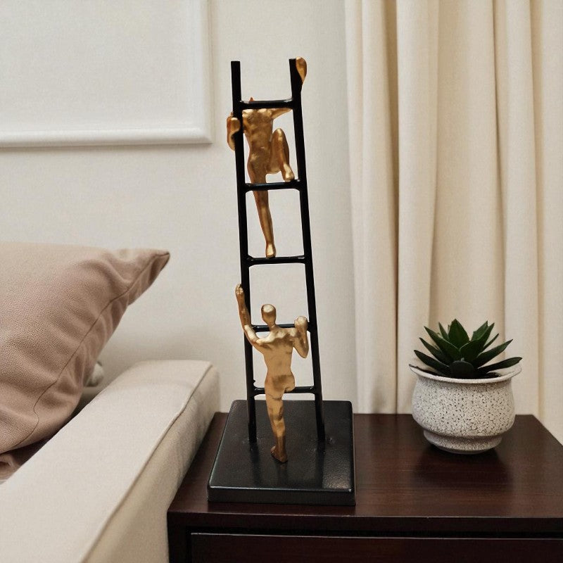 Decorative ladder sculpture with gold figures on a table next to a plant and pillow.