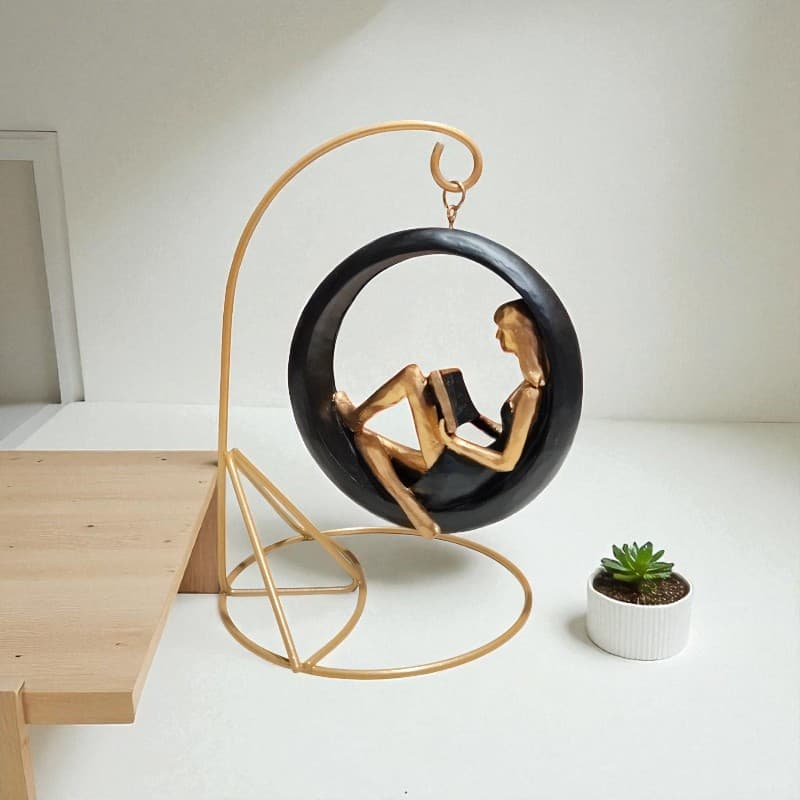 Decorative sculpture of a woman in a black hoop chair on a gold stand, with a small plant in the background.