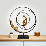 Decorative sculpture with two gold figures sitting on a black circular platform on a wooden surface.