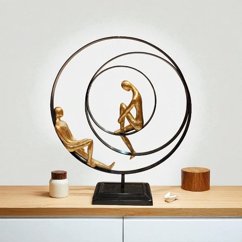 Decorative sculpture with two gold figures sitting on a black circular platform on a wooden surface.