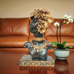 Decorative bust with floral headpiece and jewelry on a table with a plant in the background.